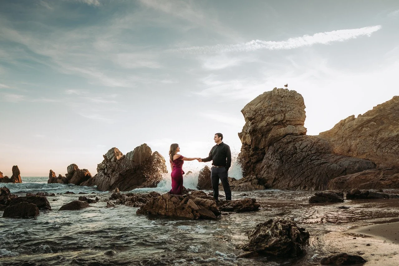 A man and woman stand on rocks in the ocean, holding hands with large rocks and a seagull in the background, during sunset.