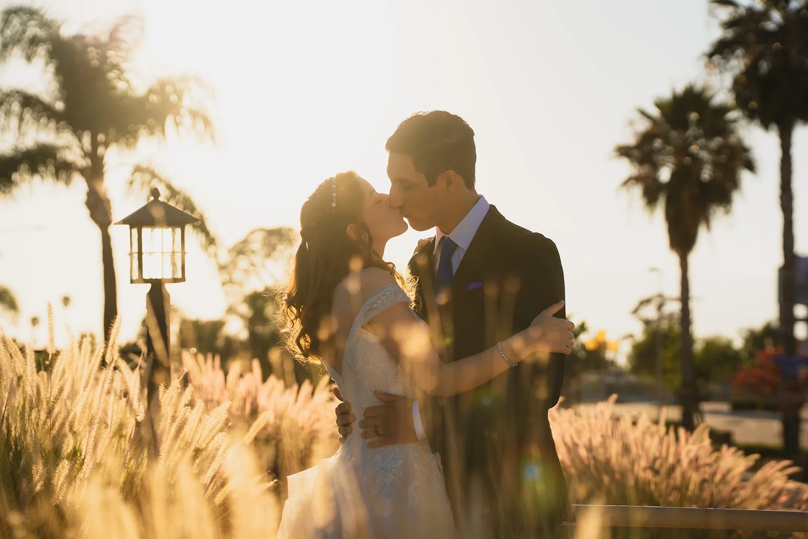 A couple kissing outdoors at sunset, with palm trees and a lamp post in the background.