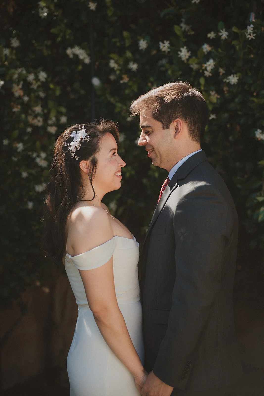 Couple on their wedding day, looking at each other and holding hands, outdoors with greenery background.
