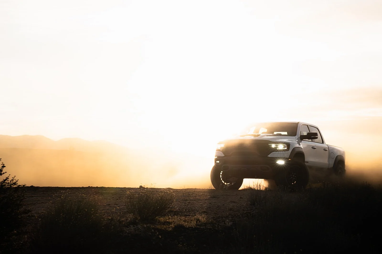 A white pickup truck driving on dirt terrain at sunset or sunrise with dust and clouds in the sky.