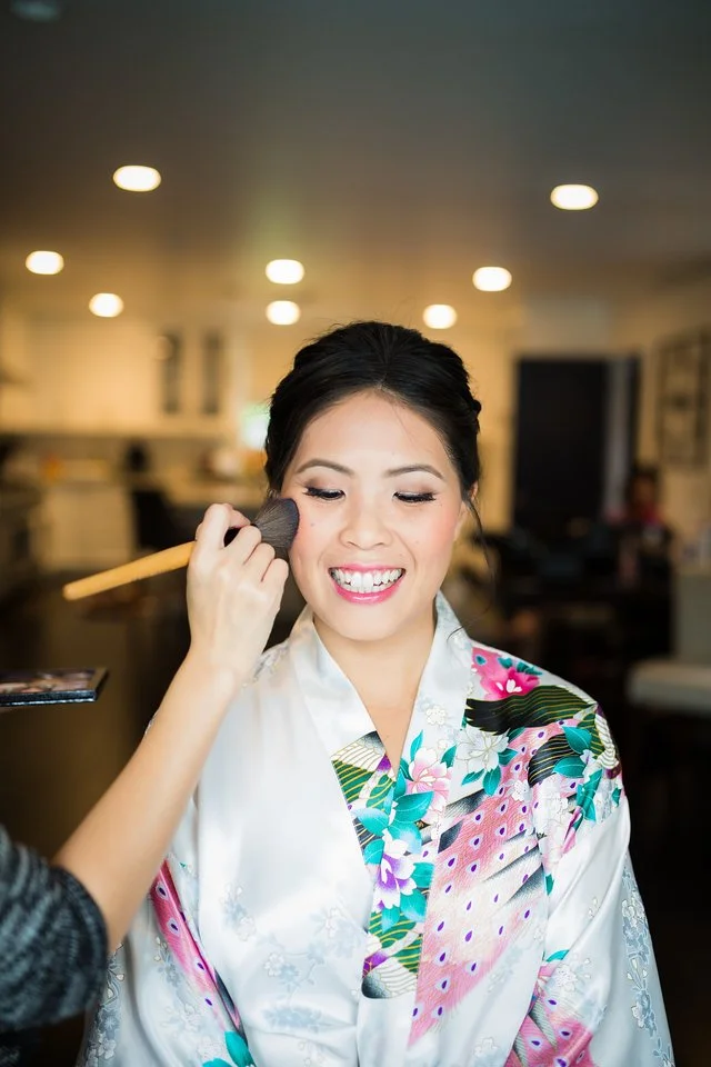 A woman wearing a colorful kimono having makeup applied with a brush in an indoor setting.