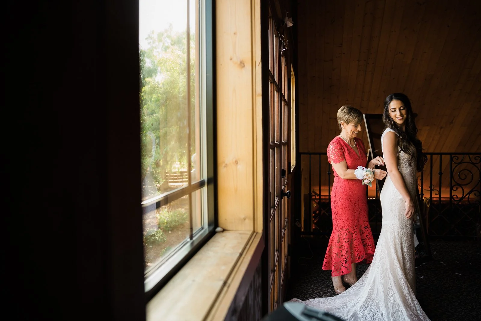 A bride in a lace wedding dress is being helped by a woman in a red lace dress, possibly her mother, inside a warmly lit room with wood-paneled walls and a large window showing a garden outside.