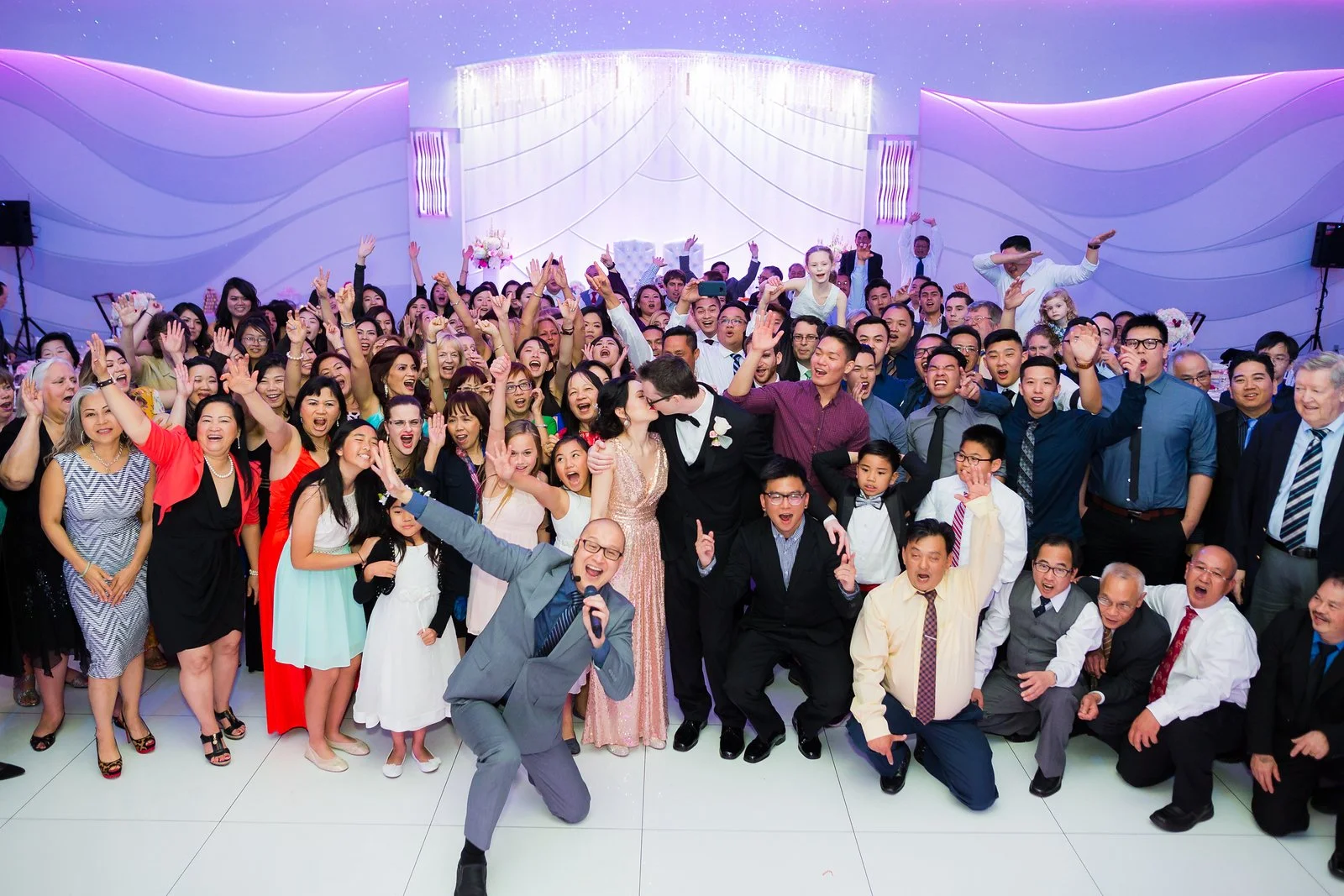 A large group of people at a celebration or wedding, with a bride and groom in the center, surrounded by friends and family. Everyone is smiling, cheering, and posing for the photo in an elegant venue with purple lighting and decorative backdrop.