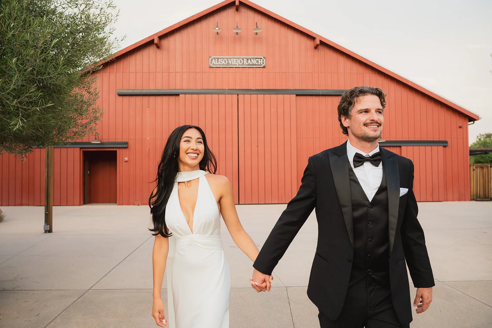 A bride and groom holding hands and walking outside in front of a red barn, smiling.