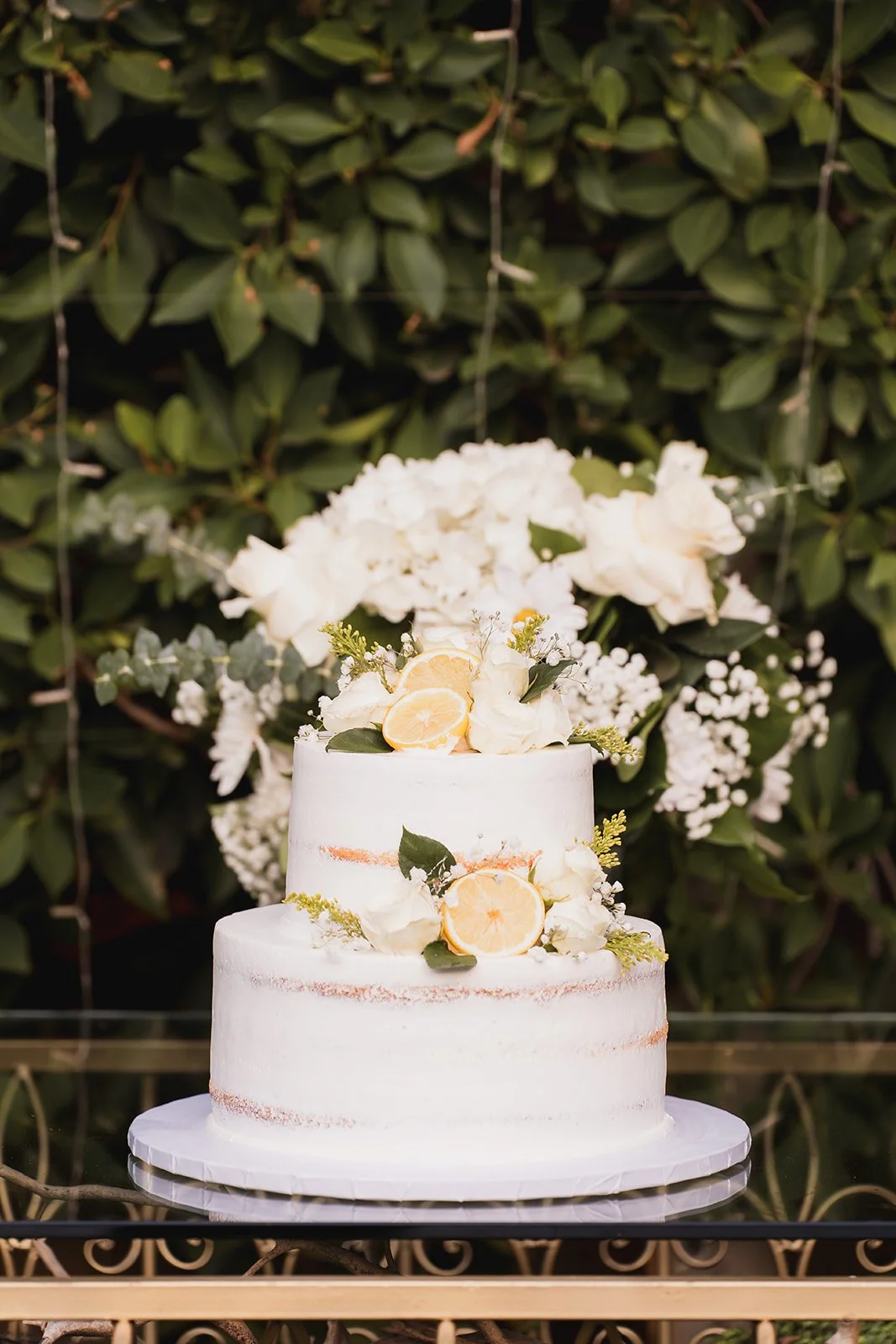 A two-tiered white wedding cake decorated with lemon slices, flowers, and greenery, set against a background of large green leaves and white flowers.