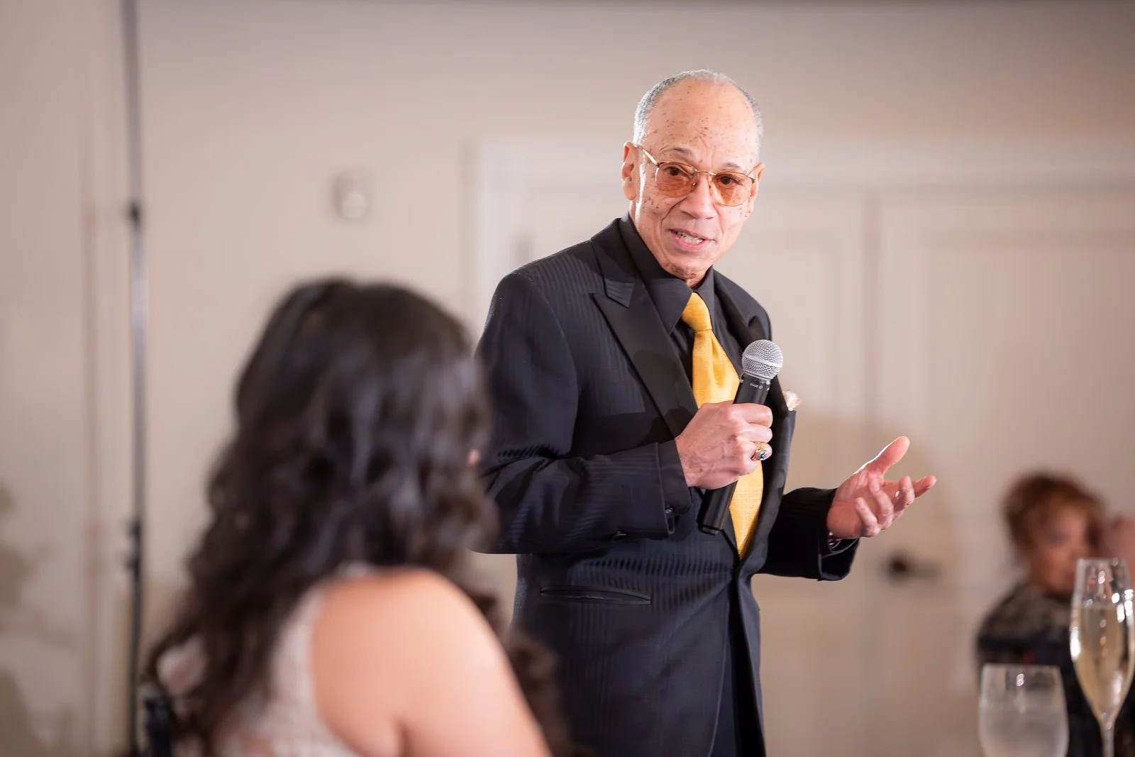 An elderly man with glasses wearing a black suit and yellow tie, holding a microphone, giving a speech at an indoor event. A woman with dark hair and a sleeveless top is seated in the foreground, with other people visible in the background.