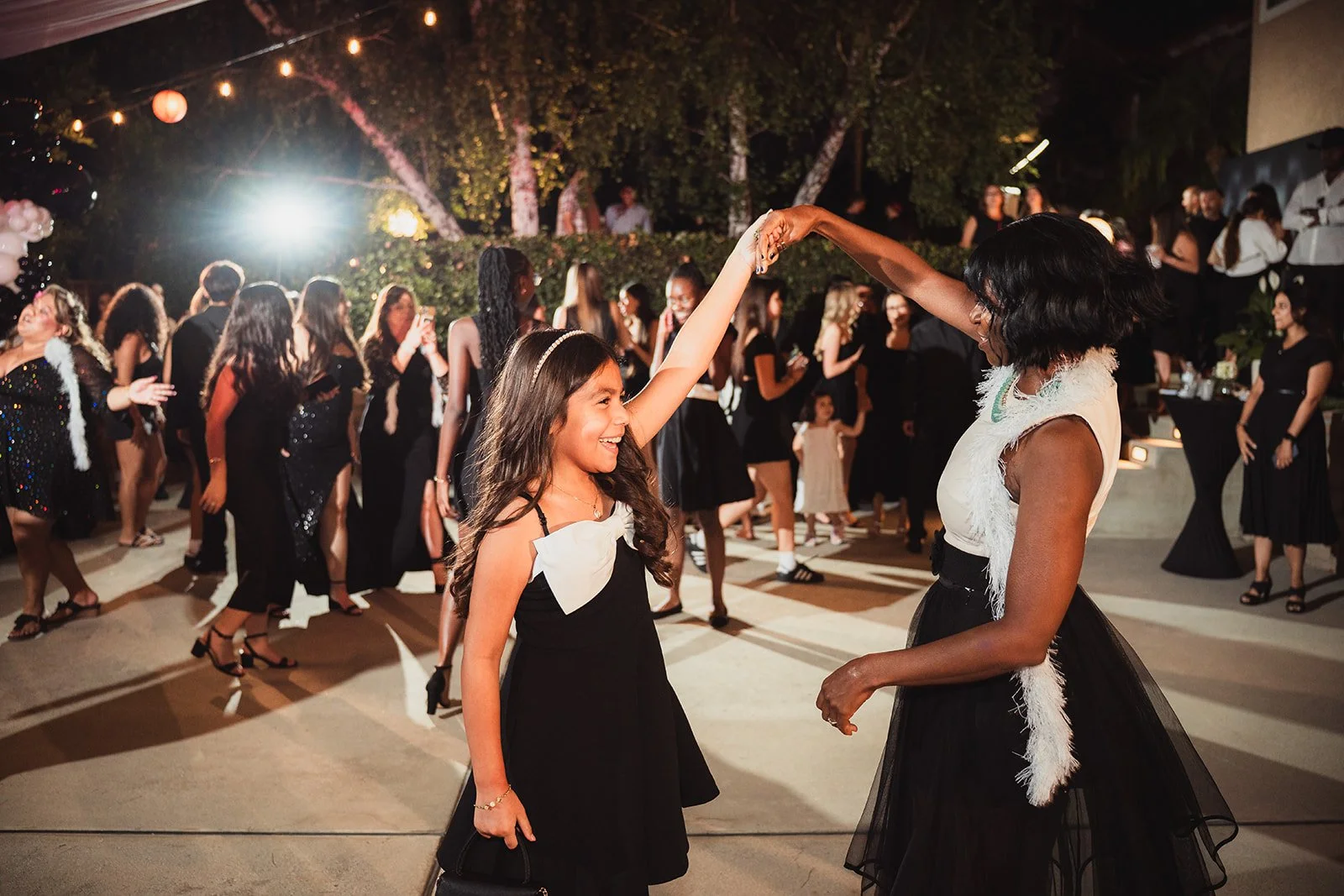 A young girl and an adult woman are dancing together at a party or celebration, surrounded by many other people dancing and socializing in the background. The scene is outdoors at night with string lights overhead.