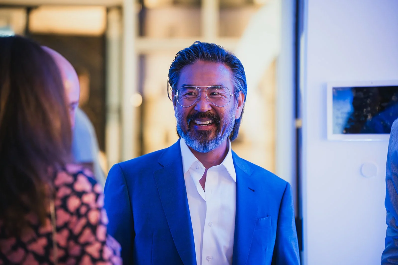 A smiling man with glasses, gray beard, and hair, dressed in a blue suit and white shirt, talking to women in a professional setting.