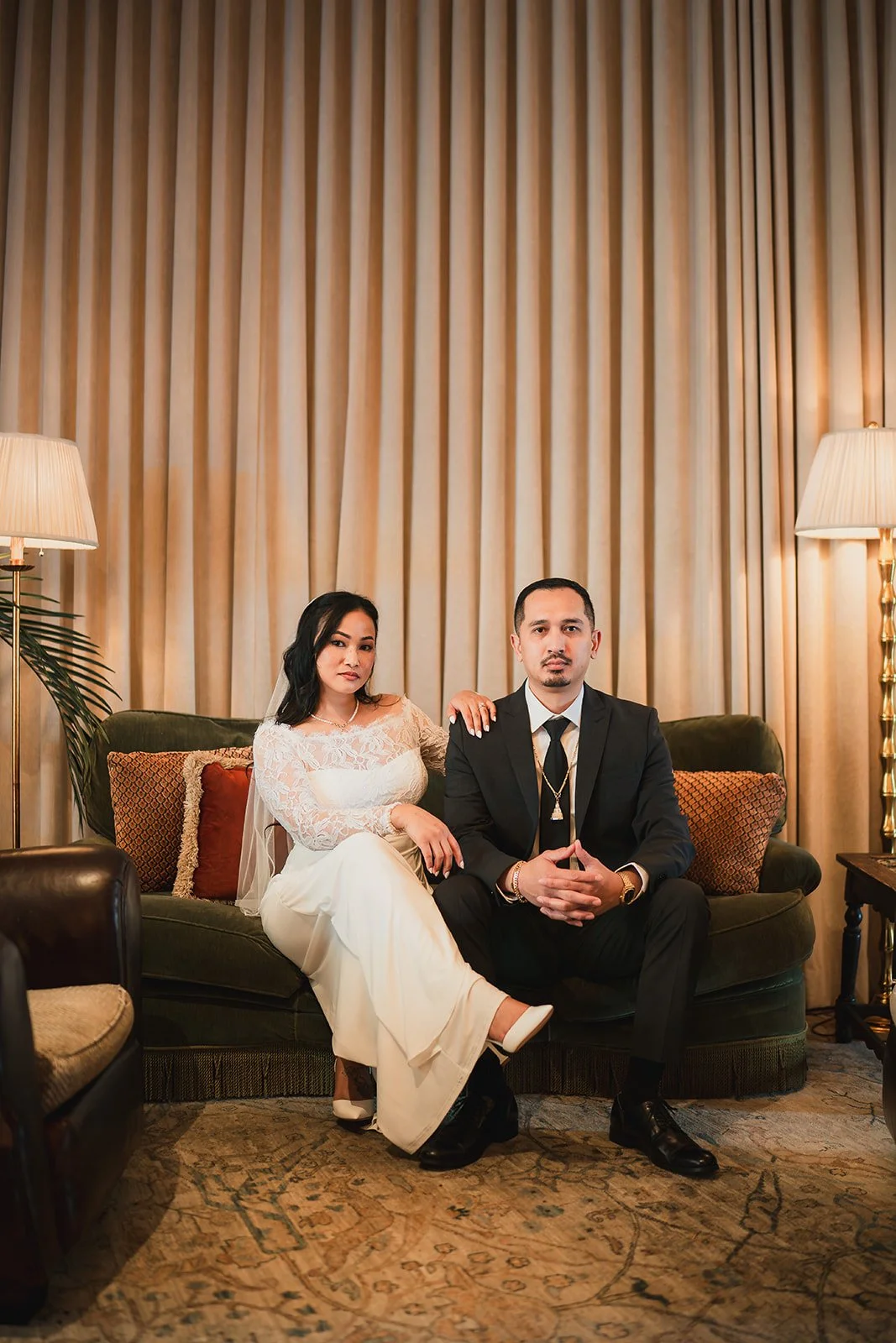 A bride and groom sitting on a green sofa in a warmly lit room with beige curtains, lamps, and decorative pillows.