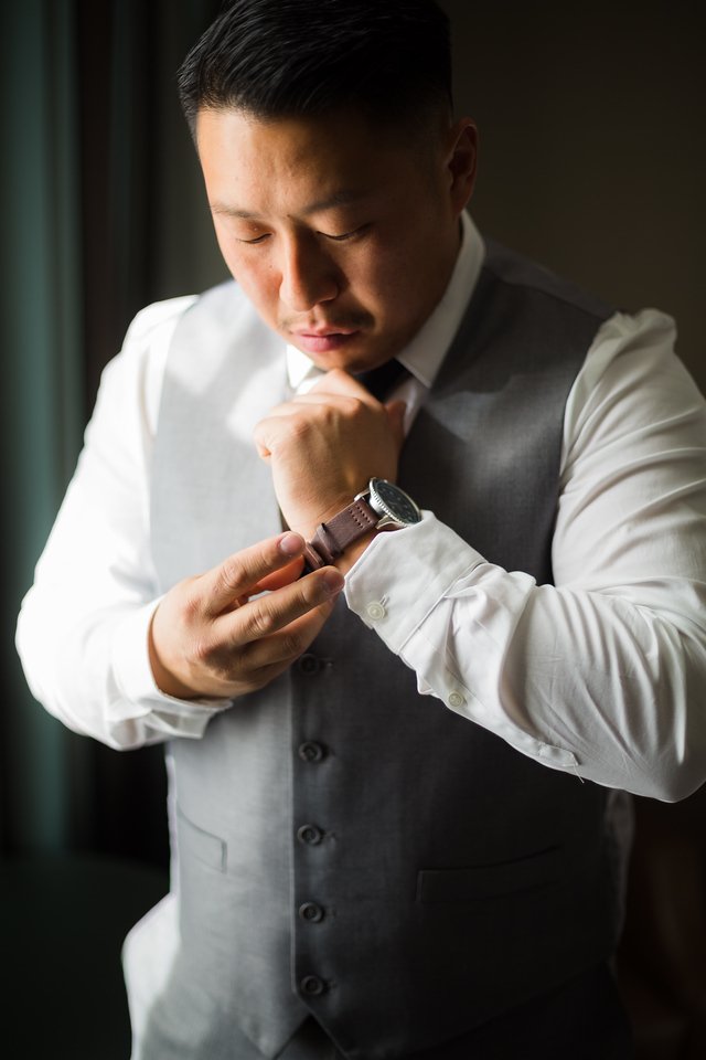 A man dressed in a white shirt and grey vest looking at his wristwatch, with one hand touching his chin, standing indoors near a window with natural light.