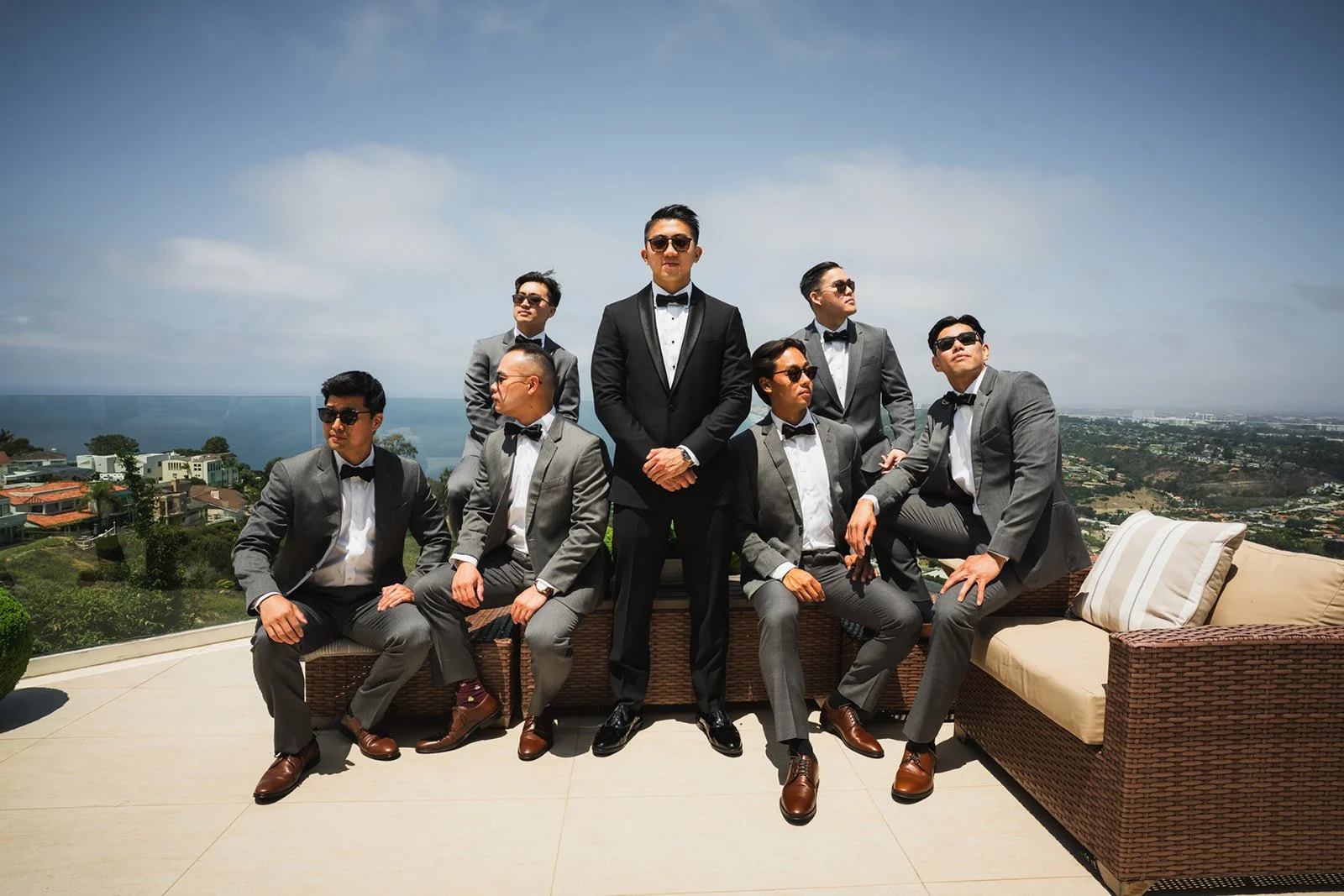 A group of nine men in suits and sunglasses posing on a balcony overlooking a city and ocean. One man stands at the center, with the others seated or standing around him.