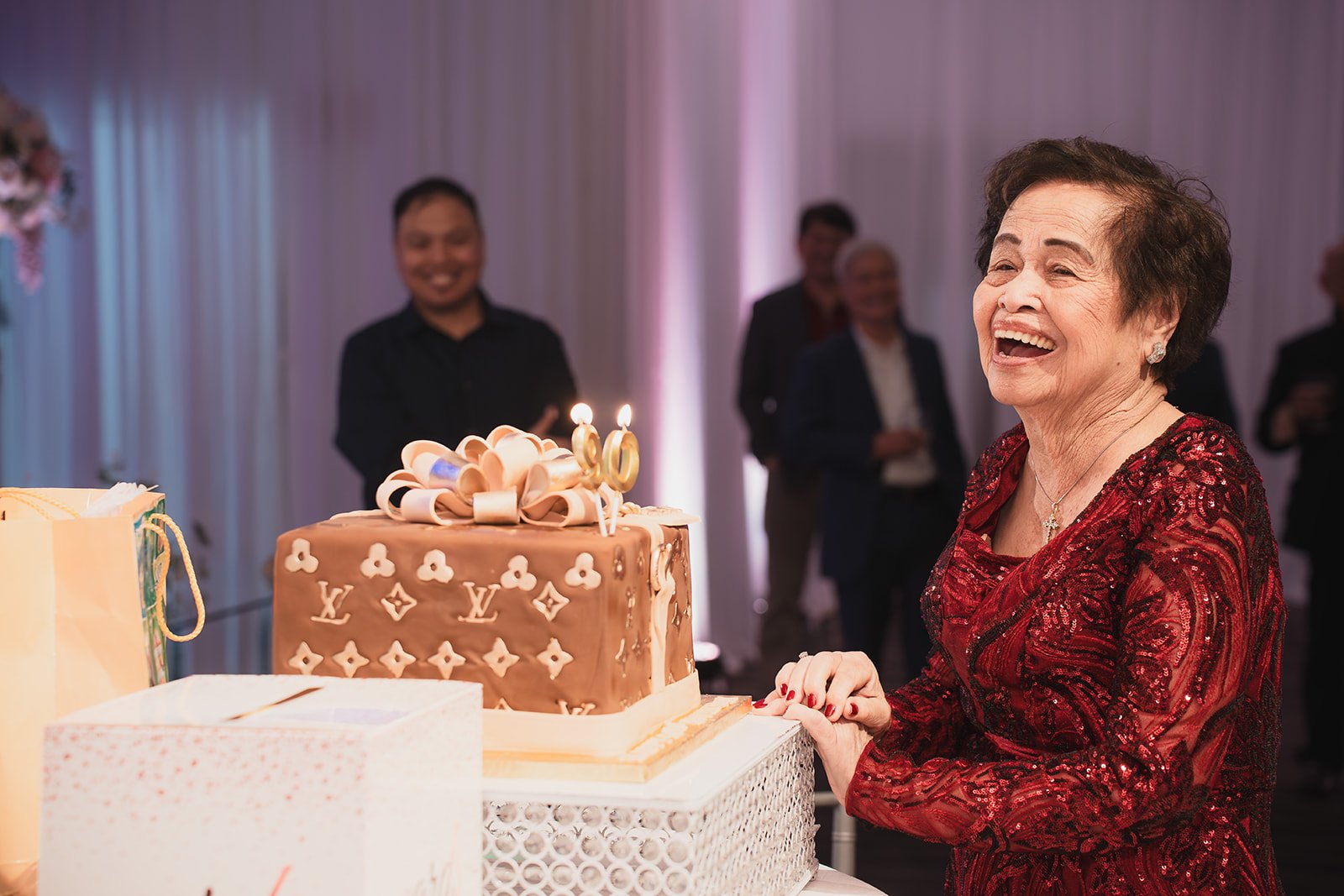 An older woman in a red dress smiling happily at a birthday celebration, standing beside a decorated birthday cake with candles, surrounded by people in a festive setting.