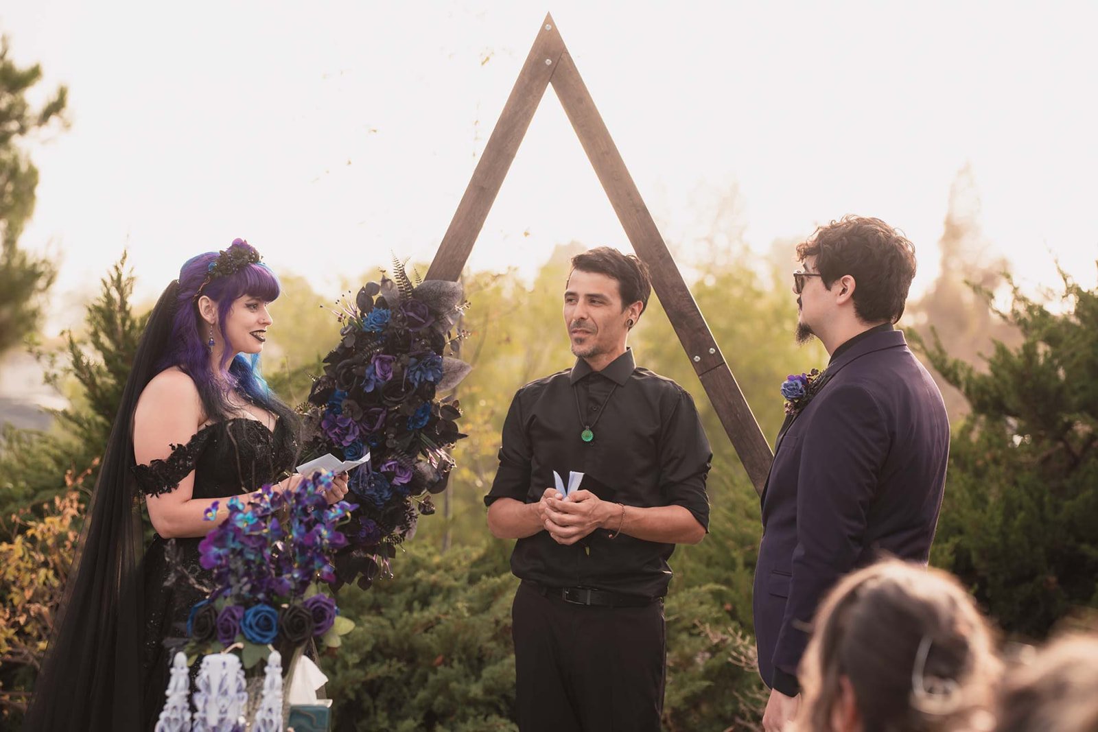 A couple in wedding attire standing before an officiant during an outdoor wedding ceremony, with a gothic style black and purple floral arrangement and a triangular wooden arch in the background.