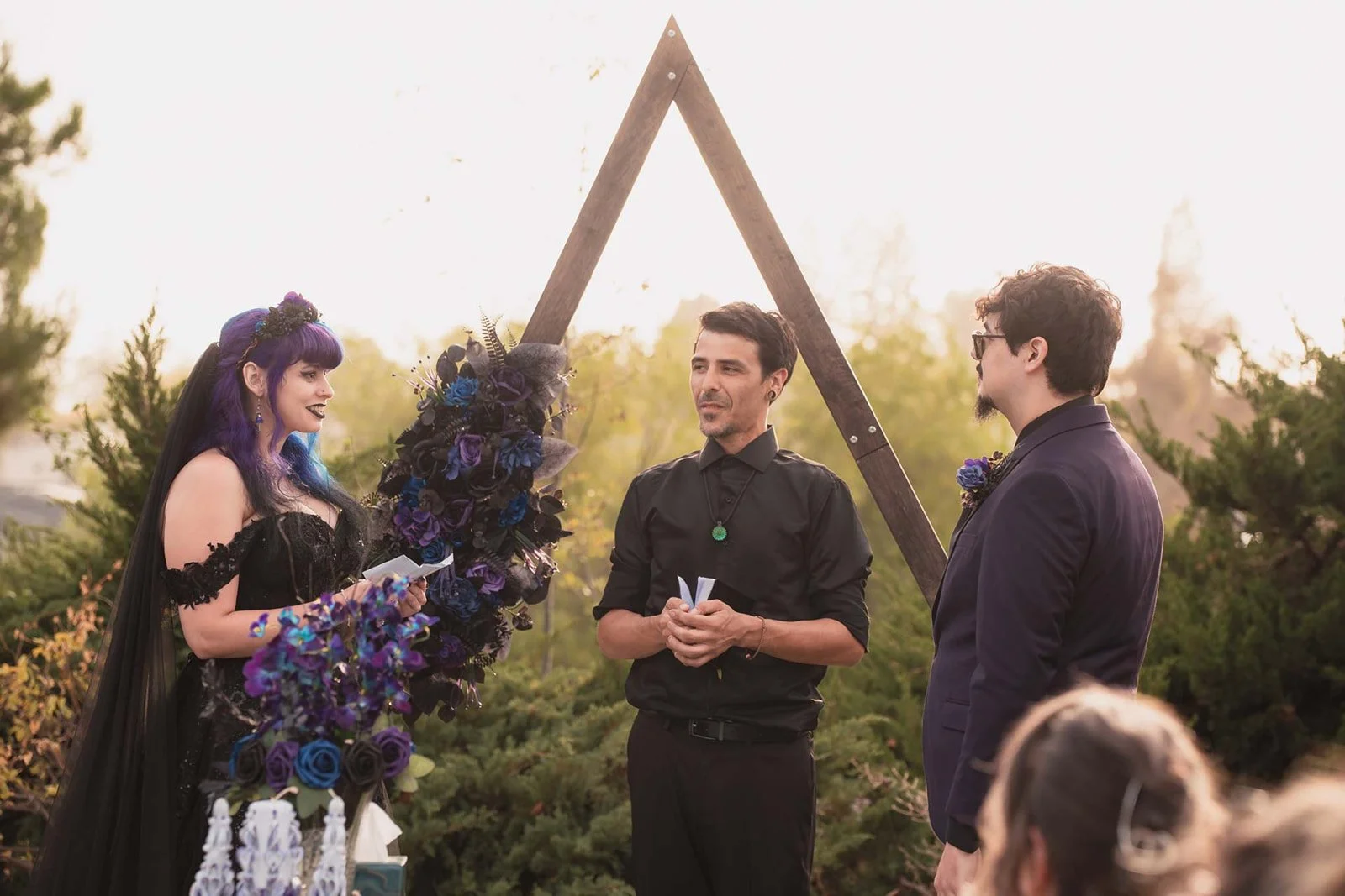 A couple in wedding attire standing before an officiant during an outdoor wedding ceremony, with a gothic style black and purple floral arrangement and a triangular wooden arch in the background.
