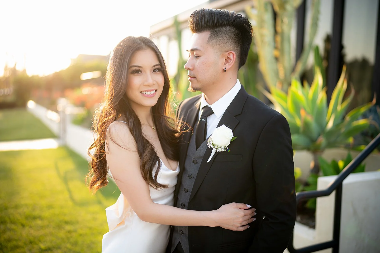 A happy couple in wedding attire standing outdoors in the sunlight. The woman has long, wavy dark hair and is smiling at the camera. The man has short, styled dark hair, is wearing a black suit with a boutonniere, and is looking at the woman. They ar