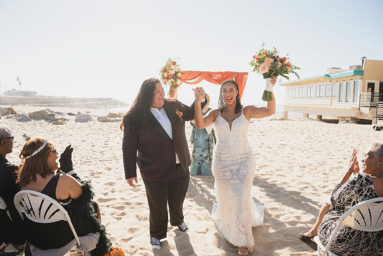 Two women, one in a wedding dress and the other in a suit, holding hands and smiling on a beach during a wedding ceremony, with guests seated nearby and a decorated arch in the background.