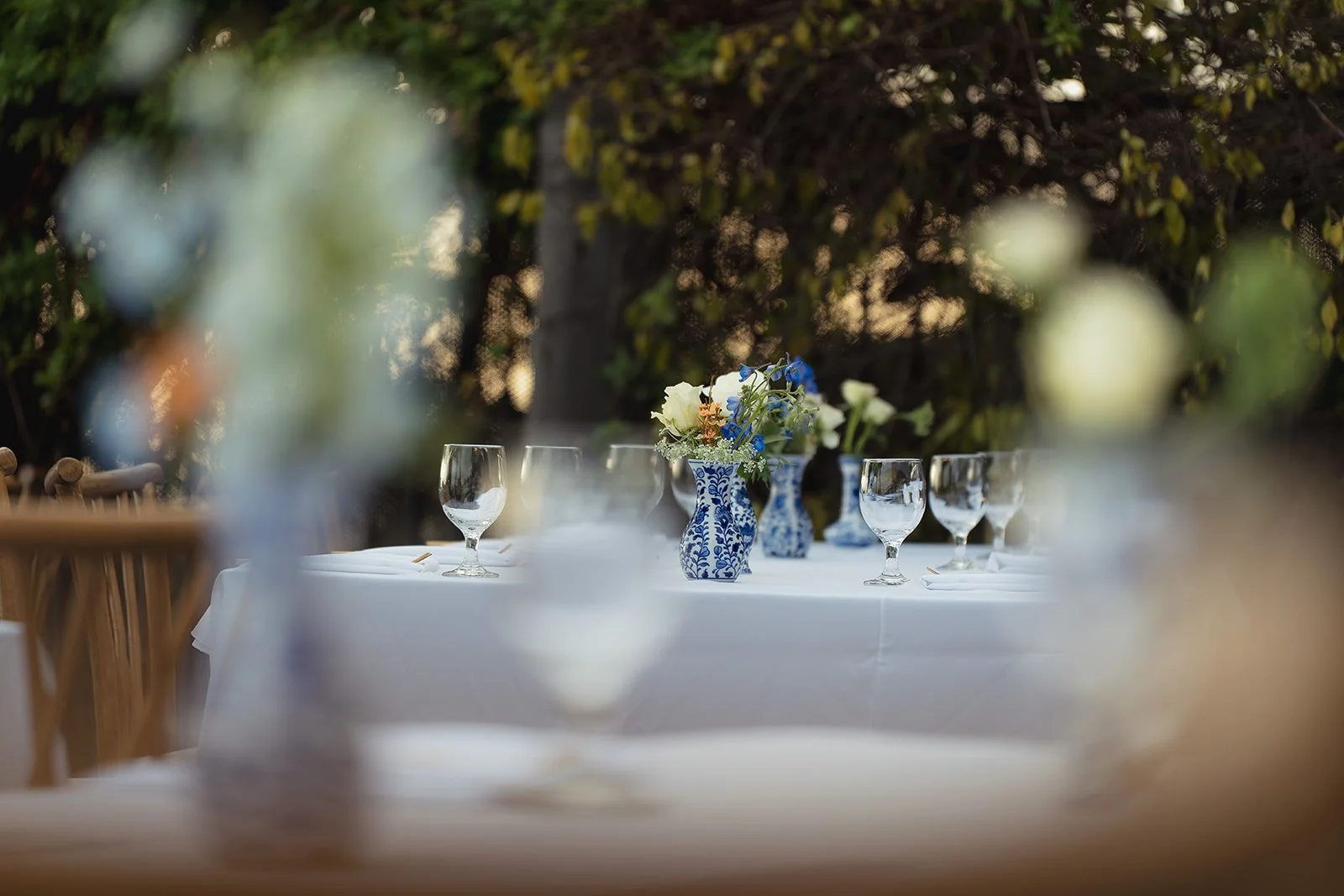 A beautifully set outdoor dining table with white tablecloth, blue and white vases with floral arrangements, and empty wine glasses. The background features greenery and plants.
