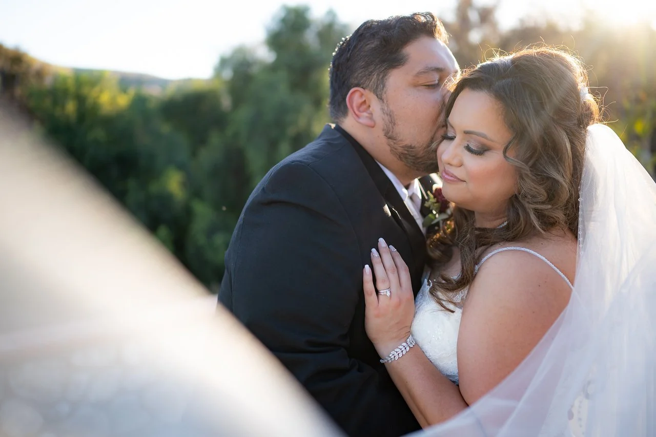 A newlywed couple sharing an intimate moment outdoors during sunset, with the groom kissing the bride on the forehead, and the bride with closed eyes and a gentle smile, wearing a white wedding dress and veil.