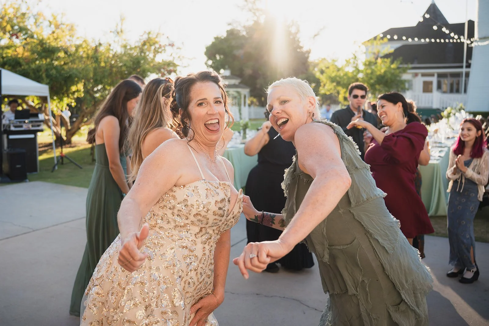 Two women dancing and smiling at an outdoor celebration during sunset, with other guests in the background.