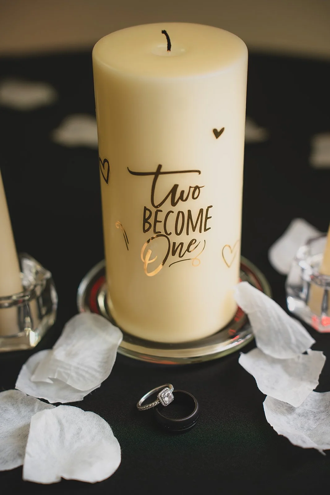 A white candle with black and gold writing that says 'two become one', surrounded by white flower petals, with an engagement ring and wedding band in front, on a black surface.
