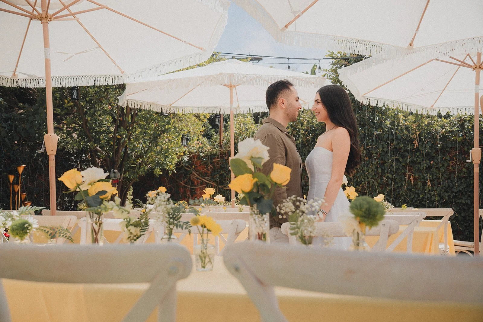 A couple standing face to face at a wedding reception outdoors, surrounded by yellow and white flowers, under white umbrellas, holding hands, looking into each other's eyes, with lush green foliage in the background.