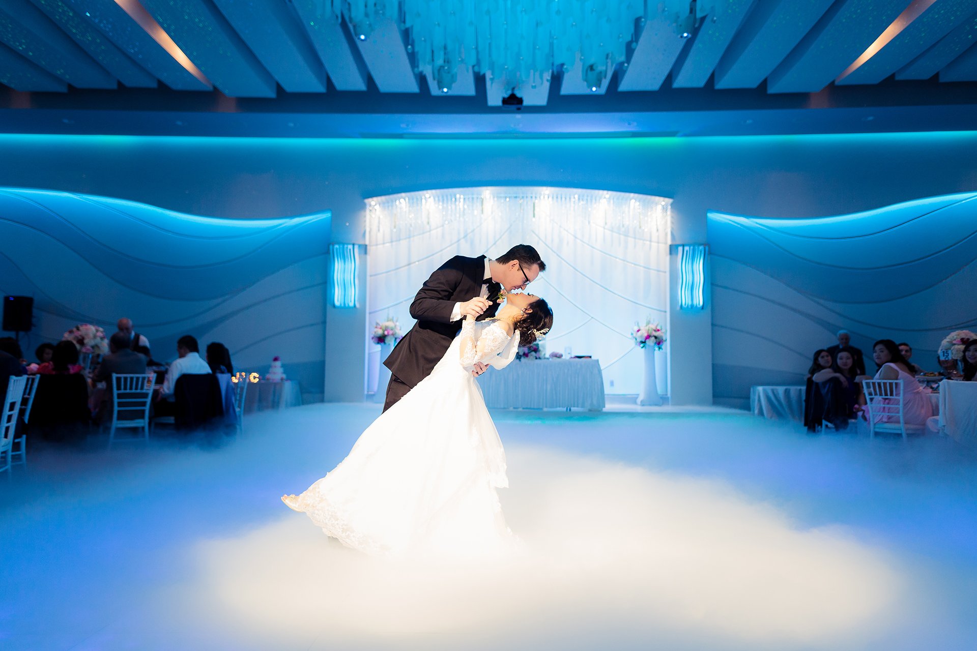 A bride and groom sharing a dance at their wedding reception, surrounded by seated guests and decorated with flowers and lighting.