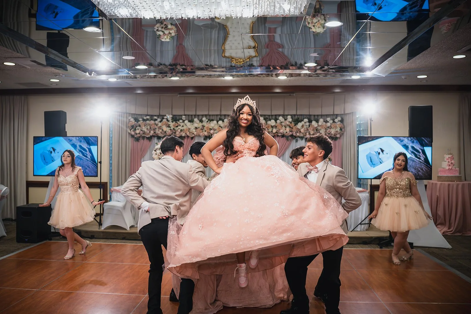A young woman in a pink ball gown with floral details, wearing a tiara, is lifted by four young men in beige suits during a celebration in a decorated banquet hall. Two young women in matching pink and gold dresses stand on either side, and the hall 