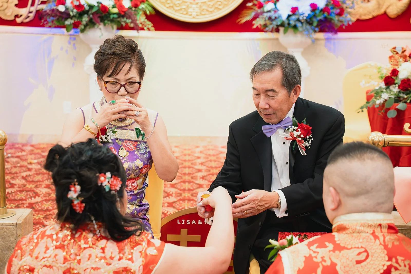 A traditional Chinese wedding ceremony with a couple, an older woman, and a young girl. The woman is dressed in traditional attire, holding a small teacup. The man is in a black tuxedo with a purple bow tie, holding the young girl's hand. The backgro