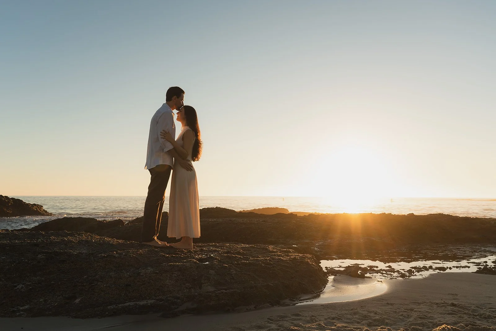 A couple standing close together on rocks at the beach during sunset, gazing into each other's eyes.