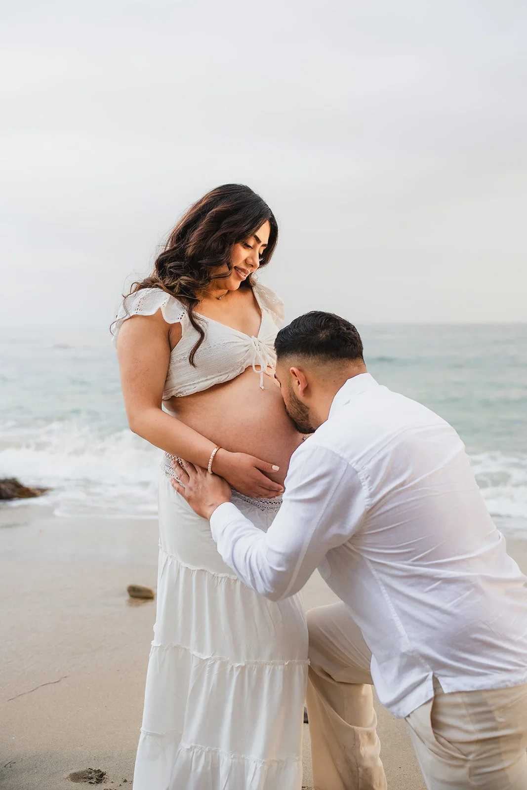 A couple on the beach during sunset, with the man kneeling and kissing the pregnant woman's belly, while she smiles looking down at him.