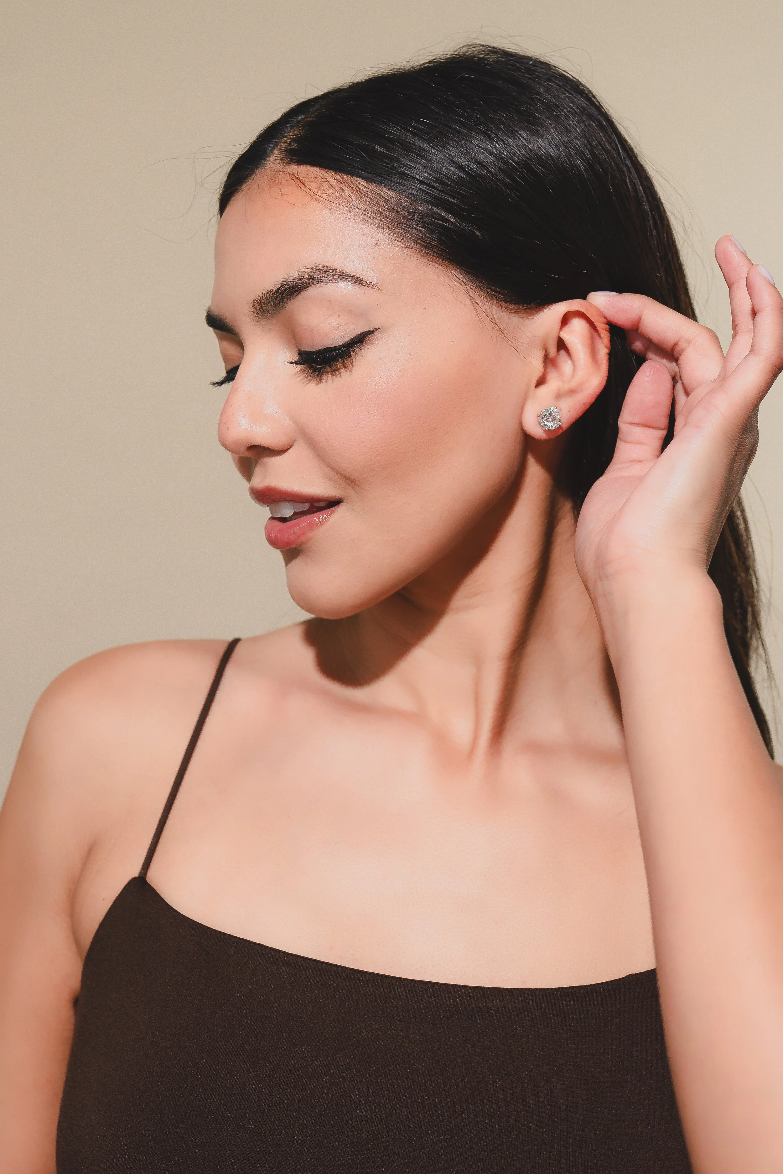 A woman with dark hair and fair skin, wearing a black spaghetti strap top, is closing her eyes and touching her ear with her left hand. She has makeup with eyeliner and glossy lips, and is wearing a large, round diamond earring.