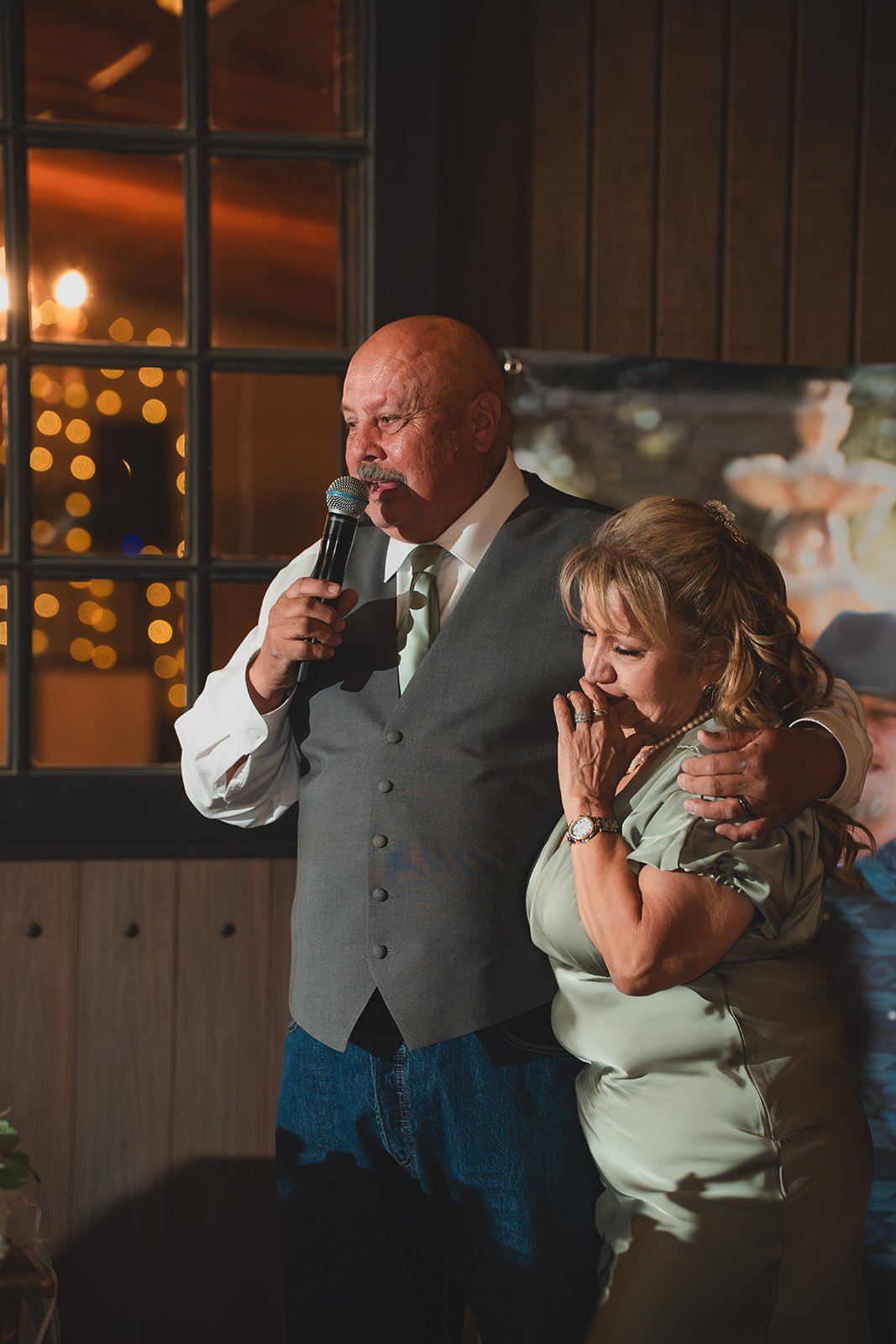 A man and woman sharing an emotional moment at a celebration; the man is speaking into a microphone and has his arm around the woman, who appears to be crying.