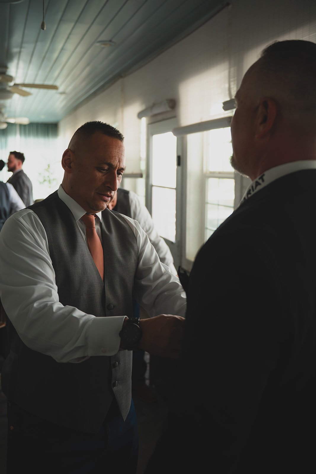 Two men dressed in formal attire, one is adjusting the other's cufflink or sleeve, inside a room with sunlight streaming through the windows. Other people are visible in the background.