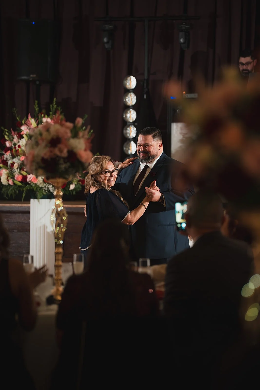A couple dancing at a wedding reception, surrounded by guests in a decorated venue with floral arrangements.