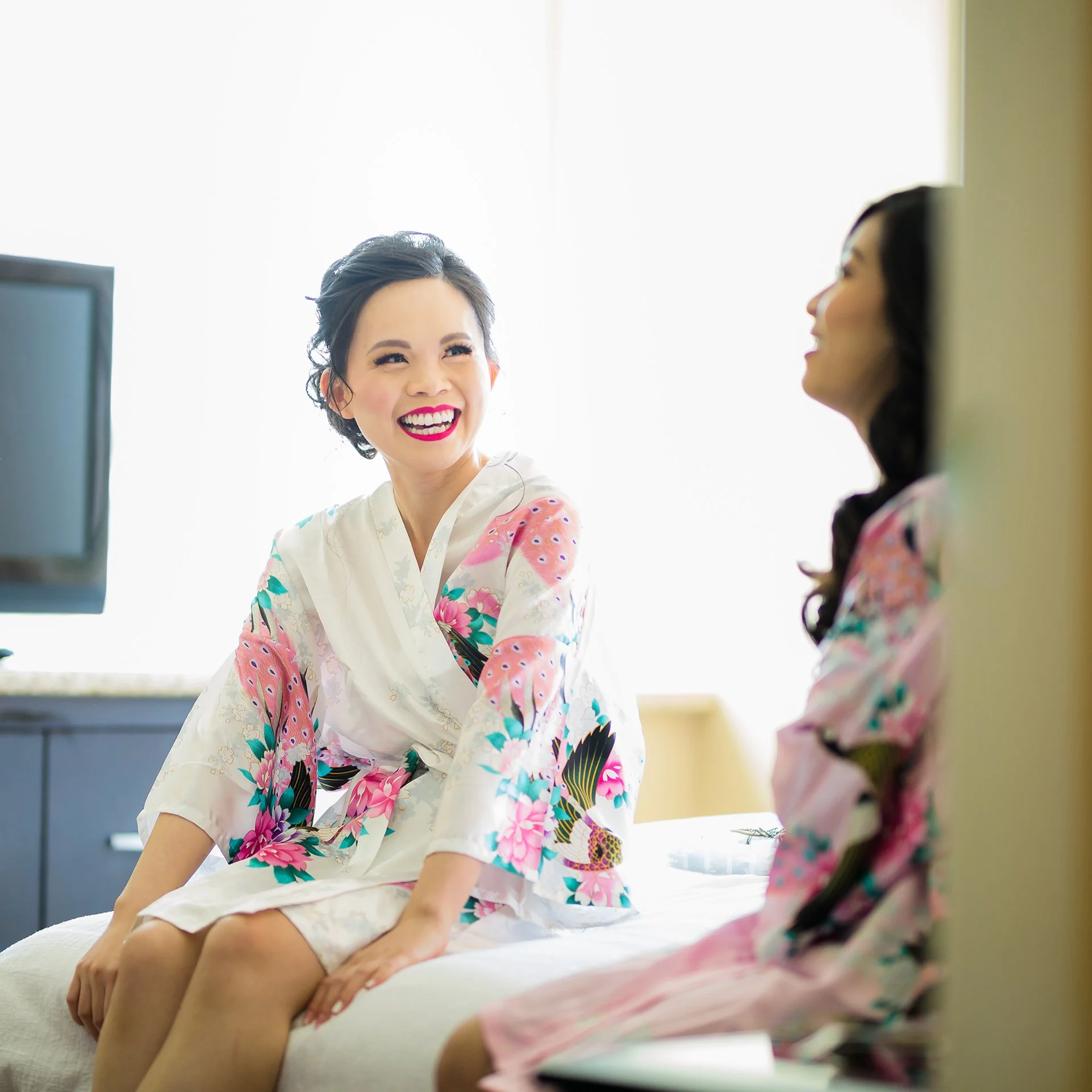 Two women in colorful floral kimonos sitting on a bed, smiling and talking in a bright room.