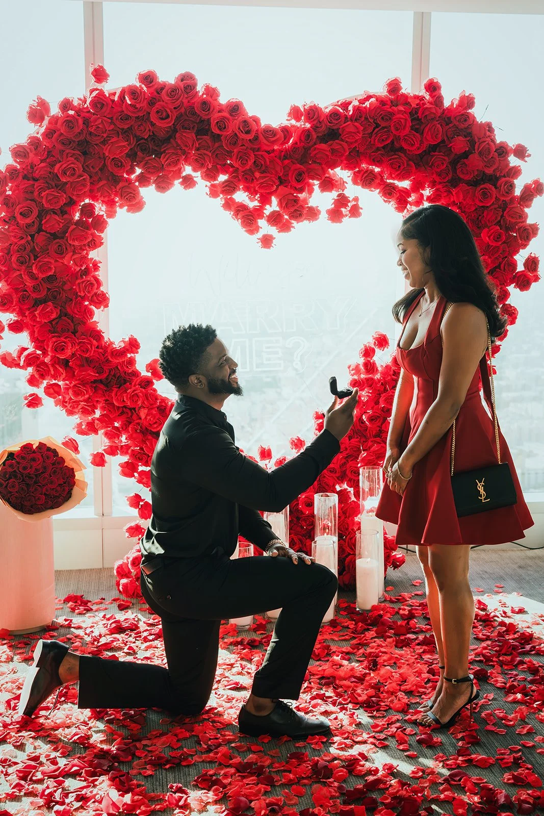 A man is kneeling on one knee and proposing marriage to a woman in front of a large heart-shaped rose arrangement. The woman is smiling and wearing a red dress, while the man is dressed in black and holding a ring box. There are rose petals scattered