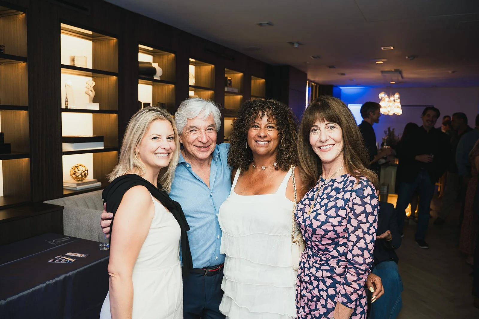 Four people standing together at an indoor social event, smiling at the camera. The group includes two women and one man, with a background of a decorated wall and other attendees mingling.