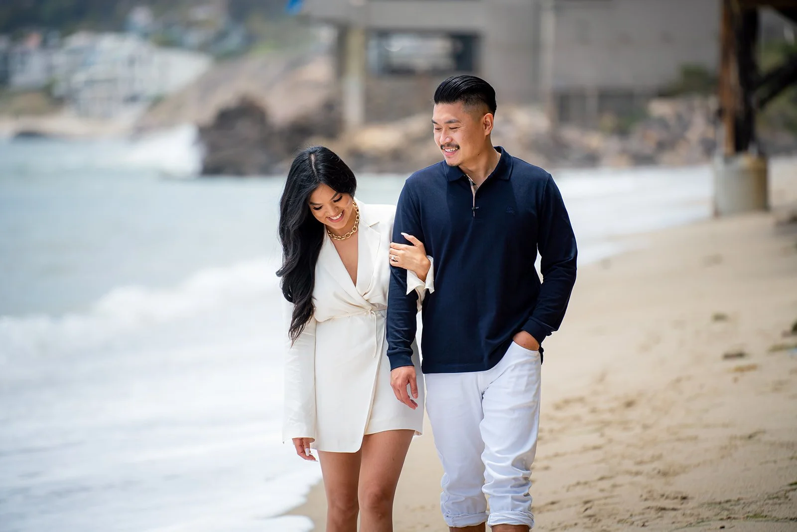 A couple walking on the beach, smiling, with the woman holding the man's arm. The woman is wearing a white dress and gold necklace, and the man is dressed in a navy blue shirt and white pants. The ocean and sandy shoreline are in the background.