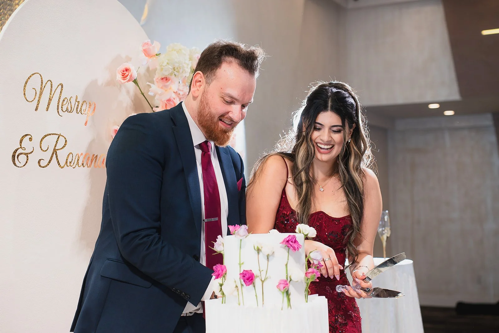 A man and woman at a wedding cake, smiling. The man wears a dark suit with a burgundy tie. The woman wears a red dress. The cake has pink and white flowers decoration. A wall with the names 'Mestrap & Rosana' and floral decorations is in the backgrou