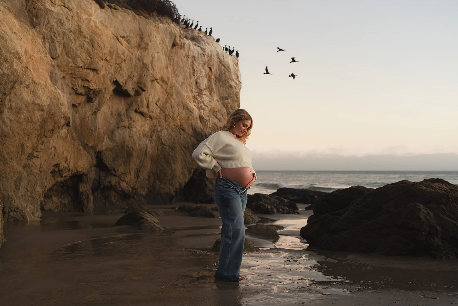 A pregnant woman stands on a beach with her hands on her belly, looking down and smiling. She is wearing a cream-colored sweater and blue jeans. Behind her, there are large rocks, a sandy shoreline, and a cliff with birds perched on top. The ocean an