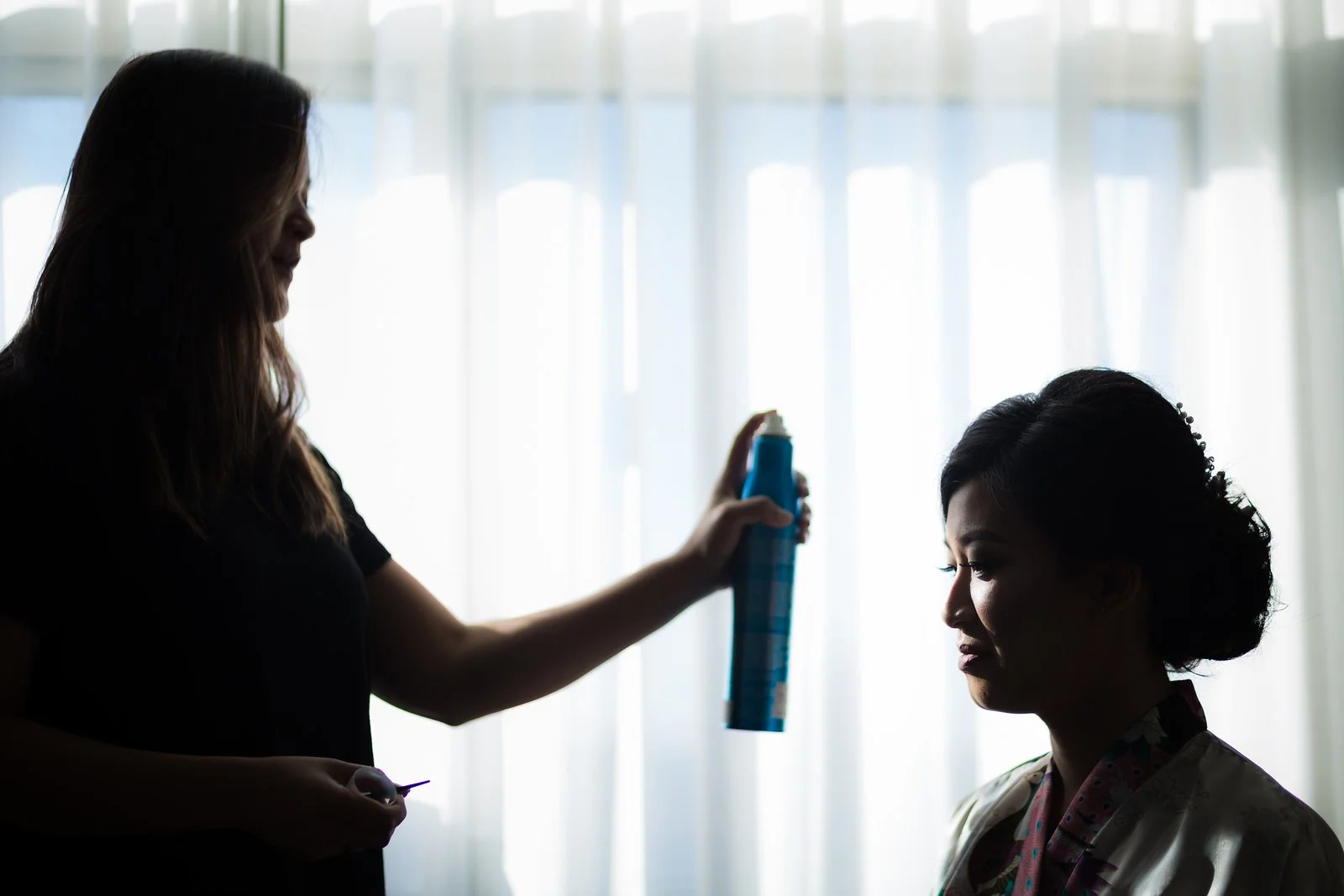 A stylist sprays hairspray onto a woman's hair during a hair styling session in a room with sheer curtains and natural light.