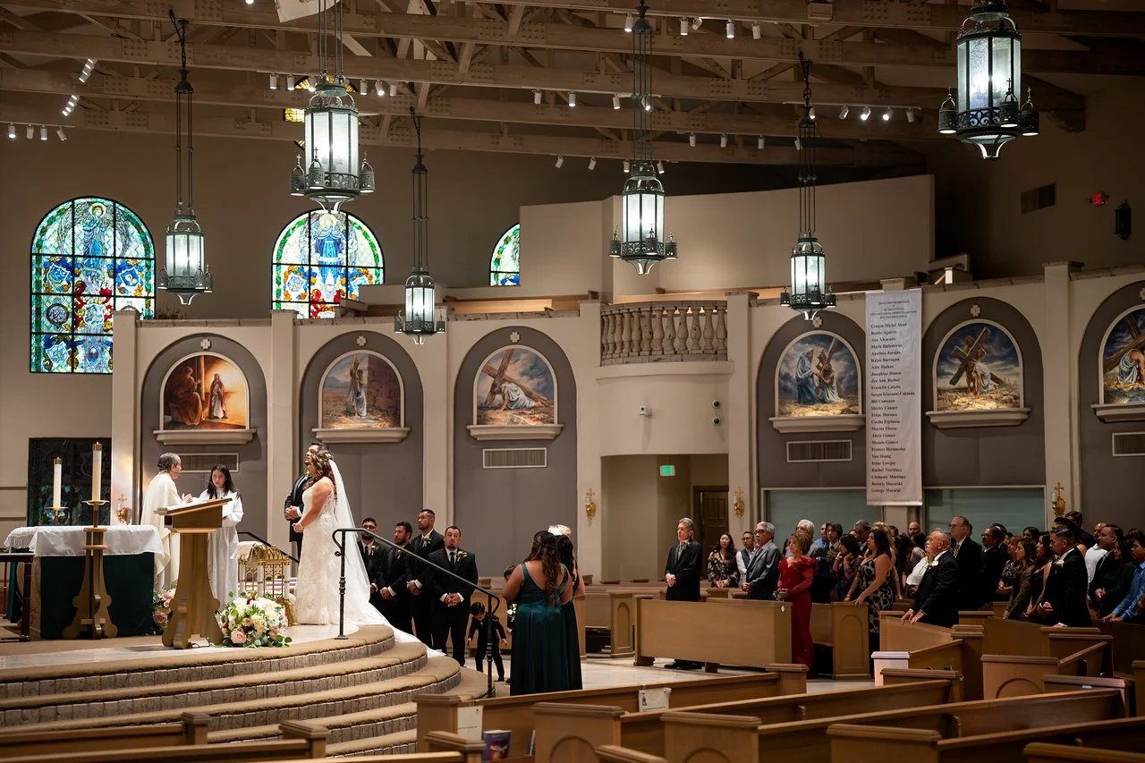 A wedding ceremony happening in a church with a bride and groom standing in front of an officiant. Guests are seated in pews, and religious artwork is displayed on the walls. Bright stained glass windows are visible above.