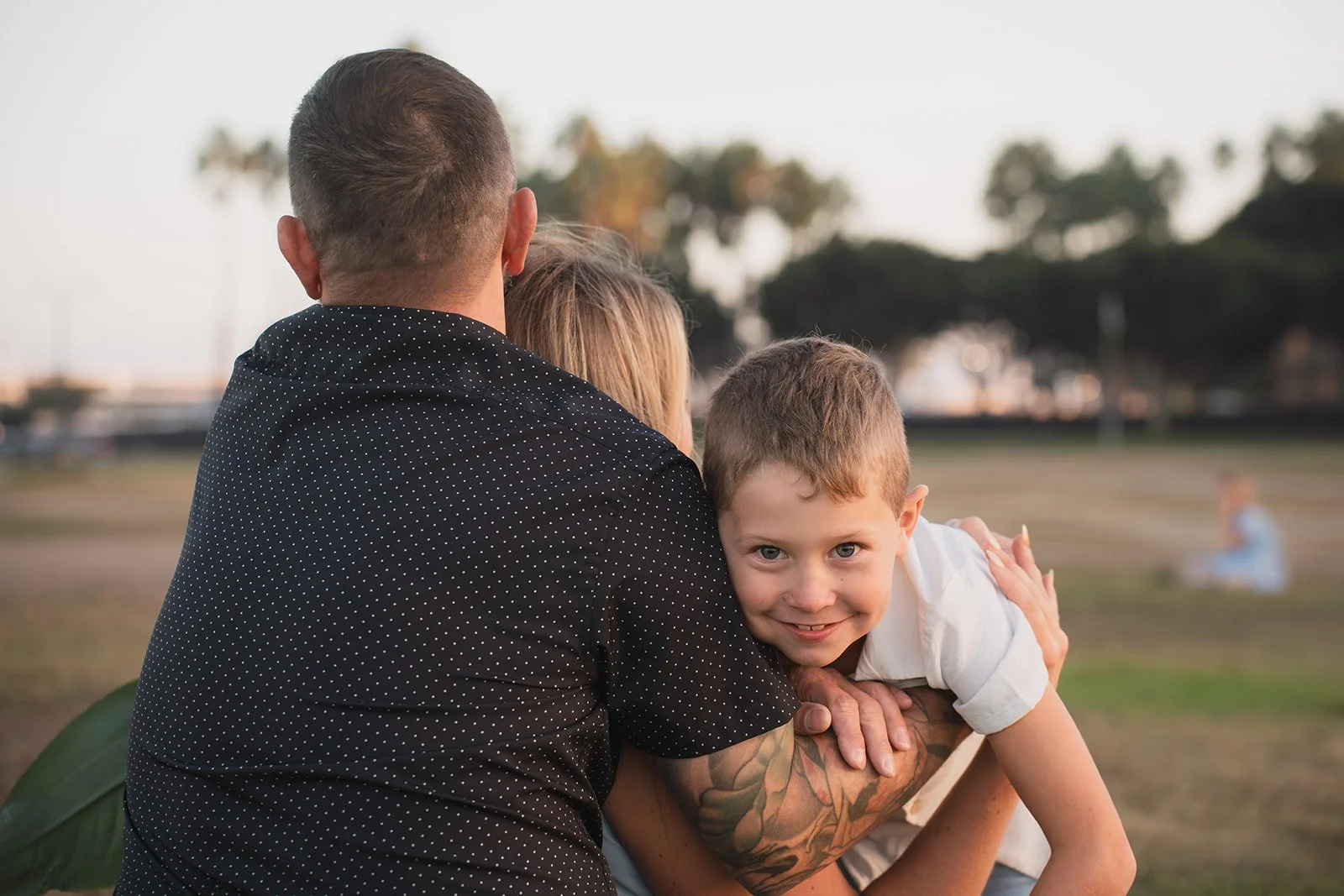 A family sitting outdoors in a park during sunset, with a young boy smiling and hugging a man, possibly his father, while another woman, possibly his mother, sits beside them.
