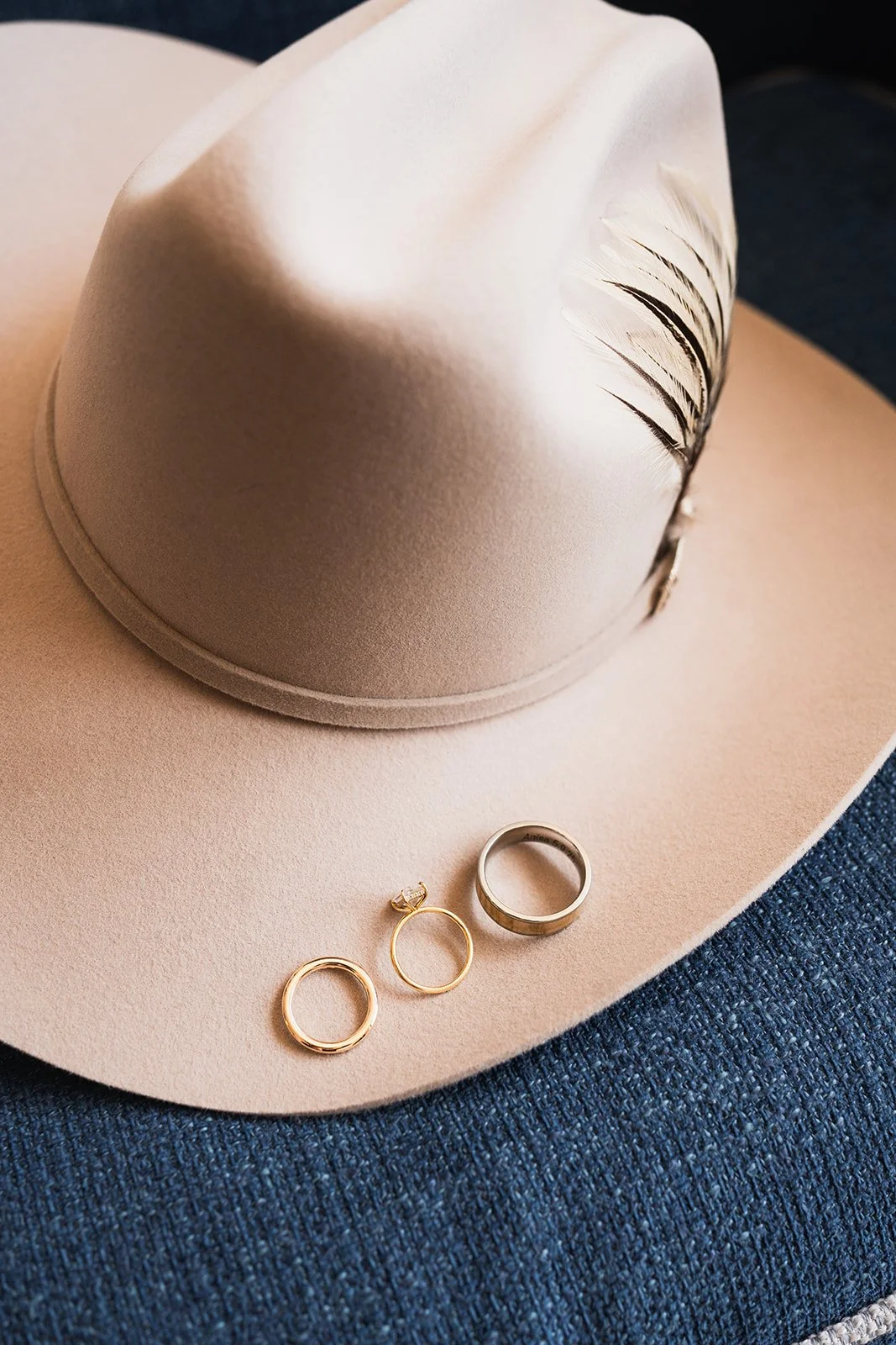 A beige cowboy hat with a wide brim and decorative feathers, along with a gold ring, a gold earring, and a silver ring on a dark blue denim fabric surface.