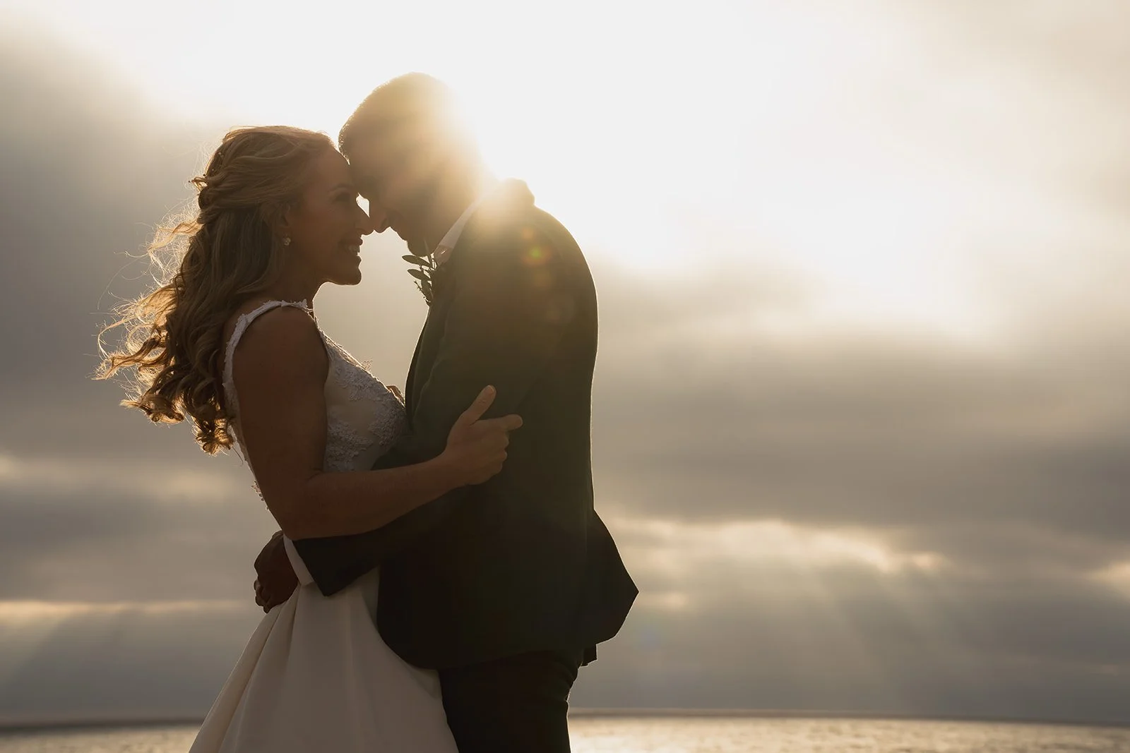 Couple dressed in wedding attire embracing on the beach with sunlight behind them and ocean in the background.