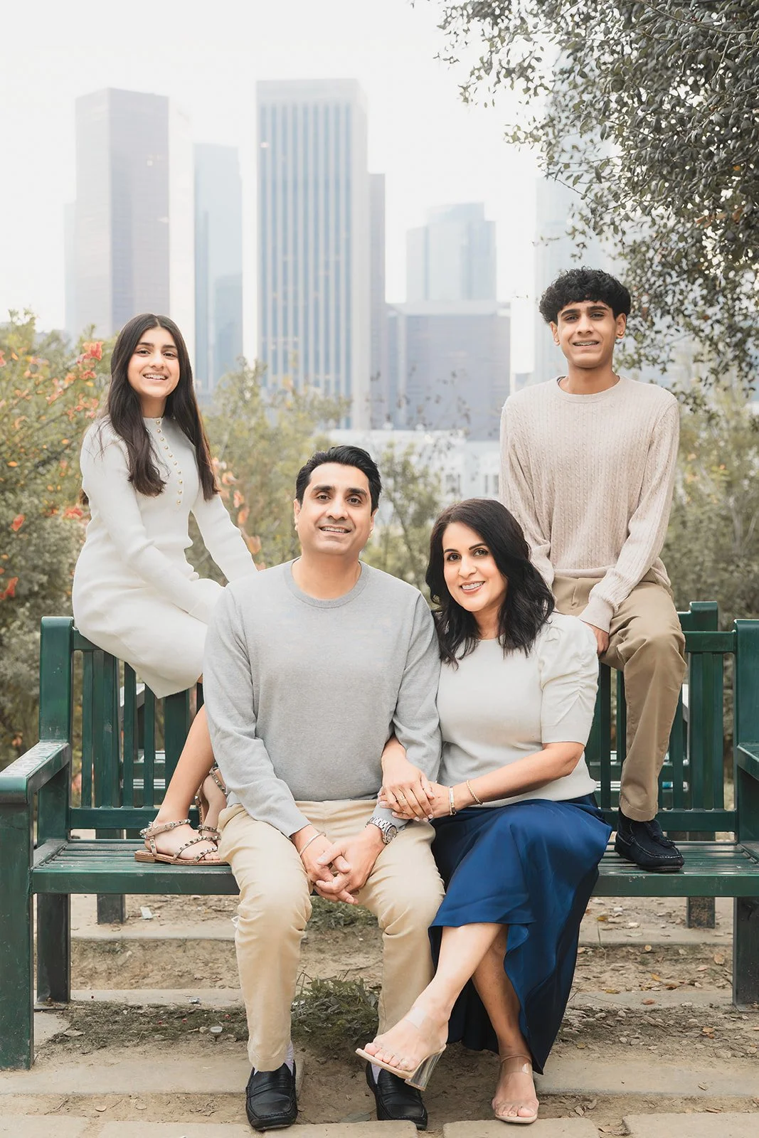 Family of four sitting on a park bench outdoors with city skyscrapers in the background, smiling at the camera.