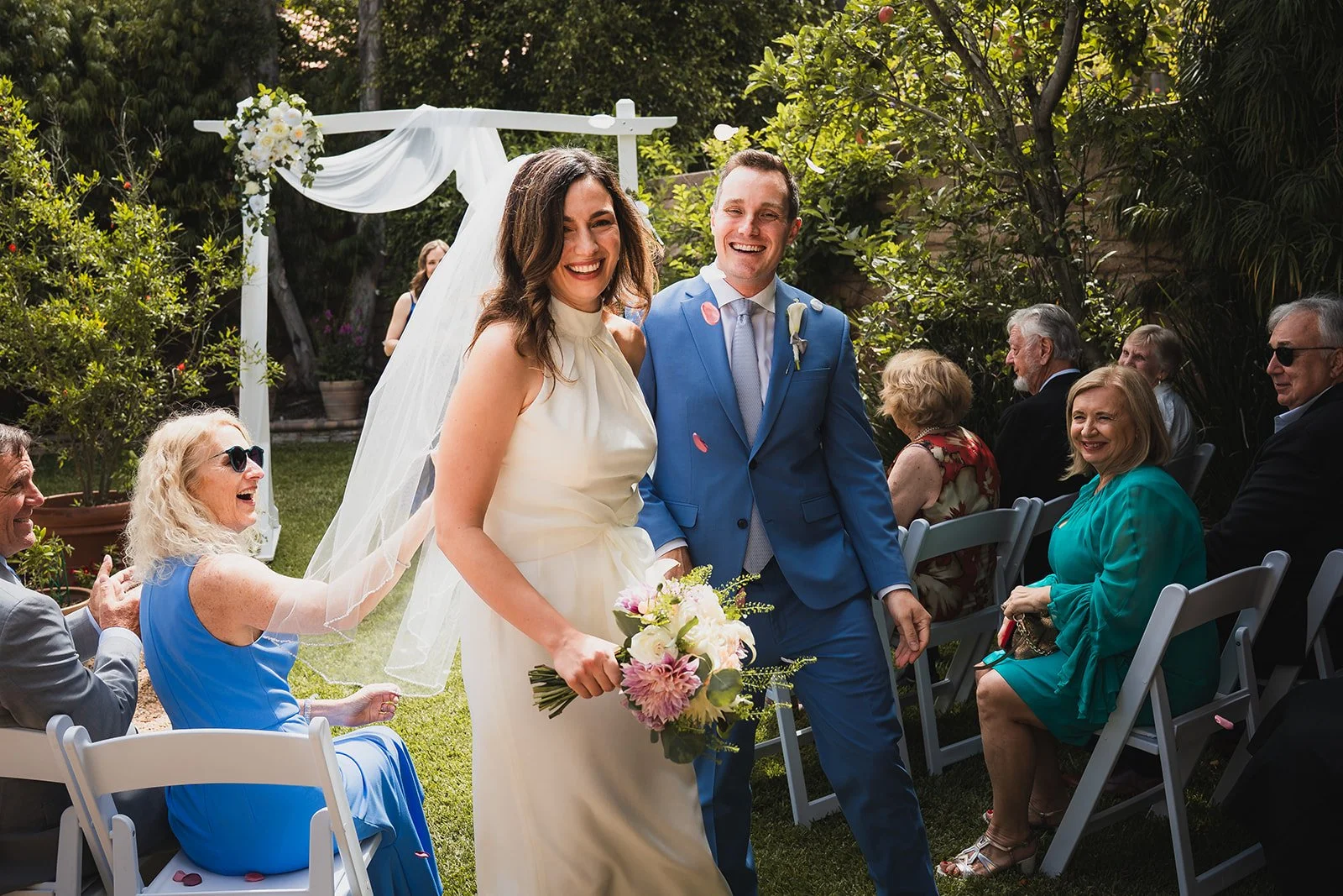 Bride in a white dress holding a bouquet of pink and white flowers, walking down the aisle with a groom in a blue suit at an outdoor wedding ceremony. Guests seated on either side, smiling and laughing, with greenery and a white wedding arch in the b