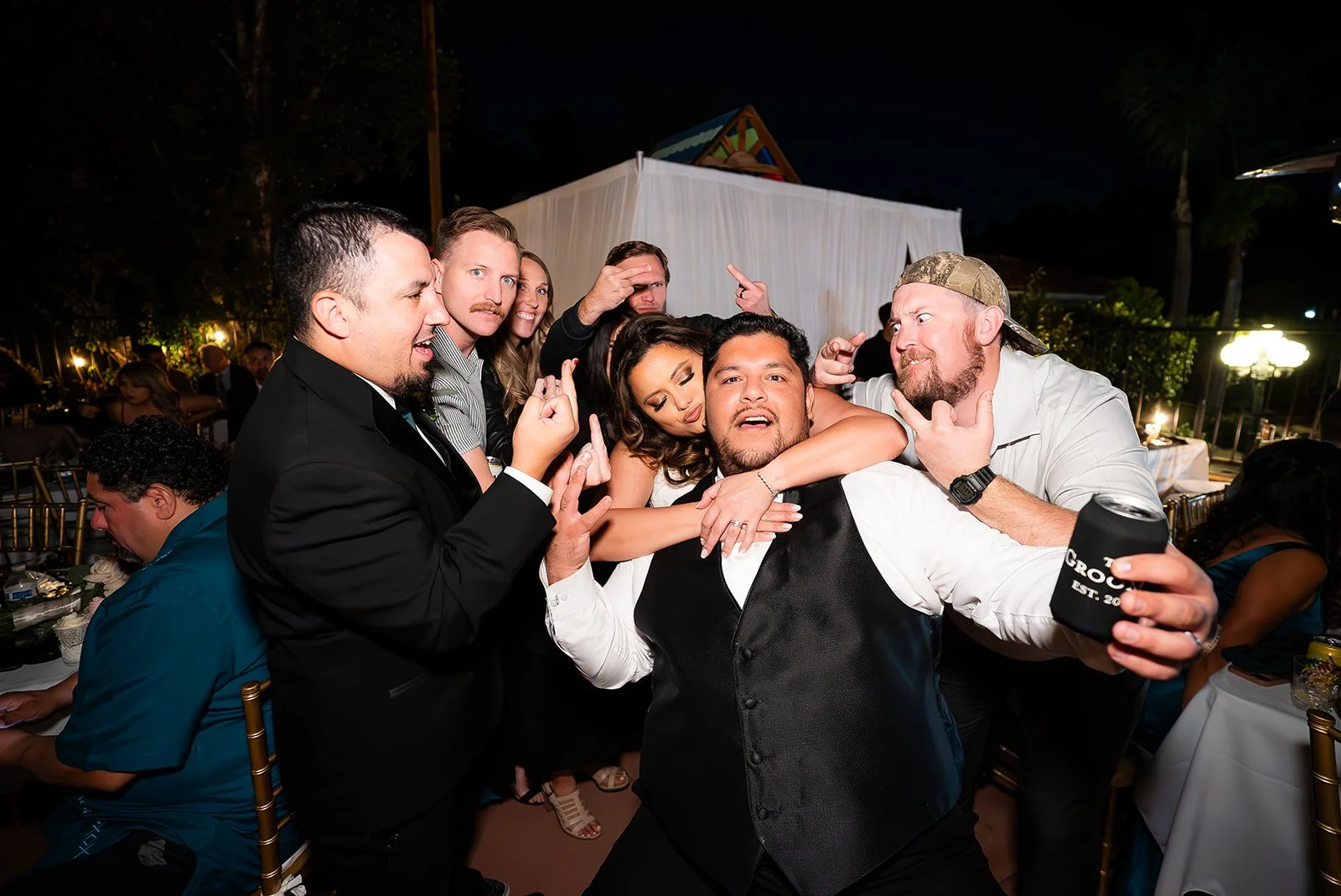 Group of people having fun at a party, with one person in a vest and others surrounding him, some making playful gestures, at nighttime outdoor event.