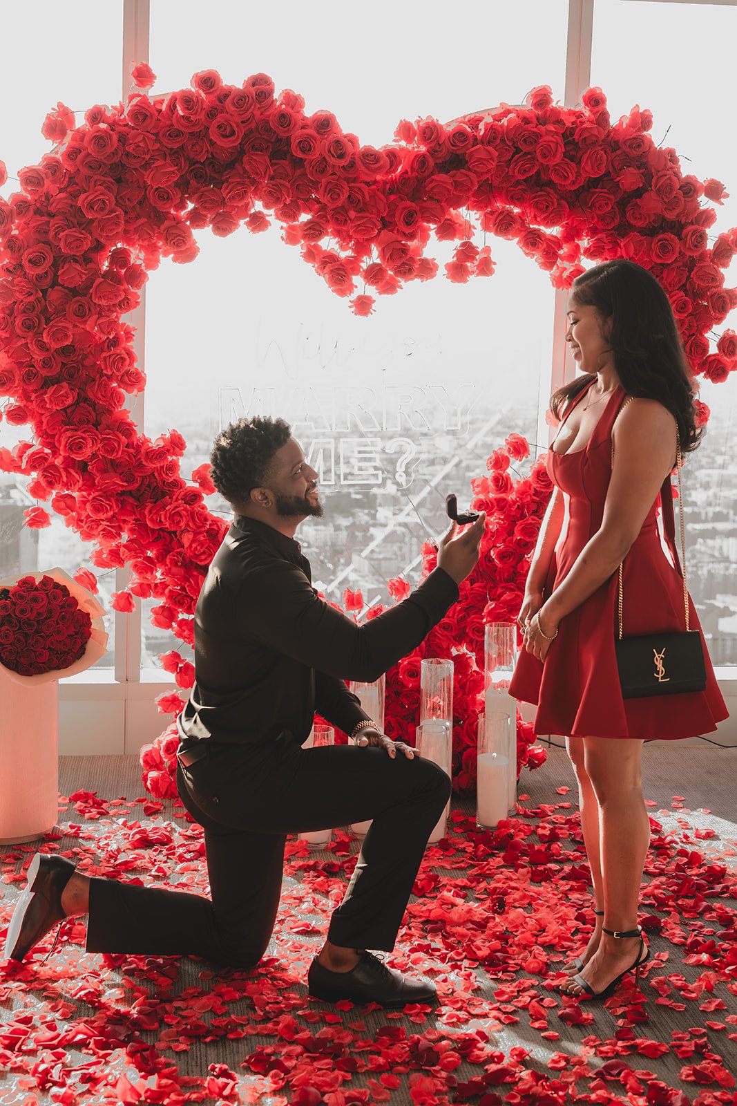 A man is kneeling on one knee and proposing marriage to a woman in front of a large heart-shaped rose arrangement. The woman is smiling and wearing a red dress, while the man is dressed in black and holding a ring box. There are rose petals scattered