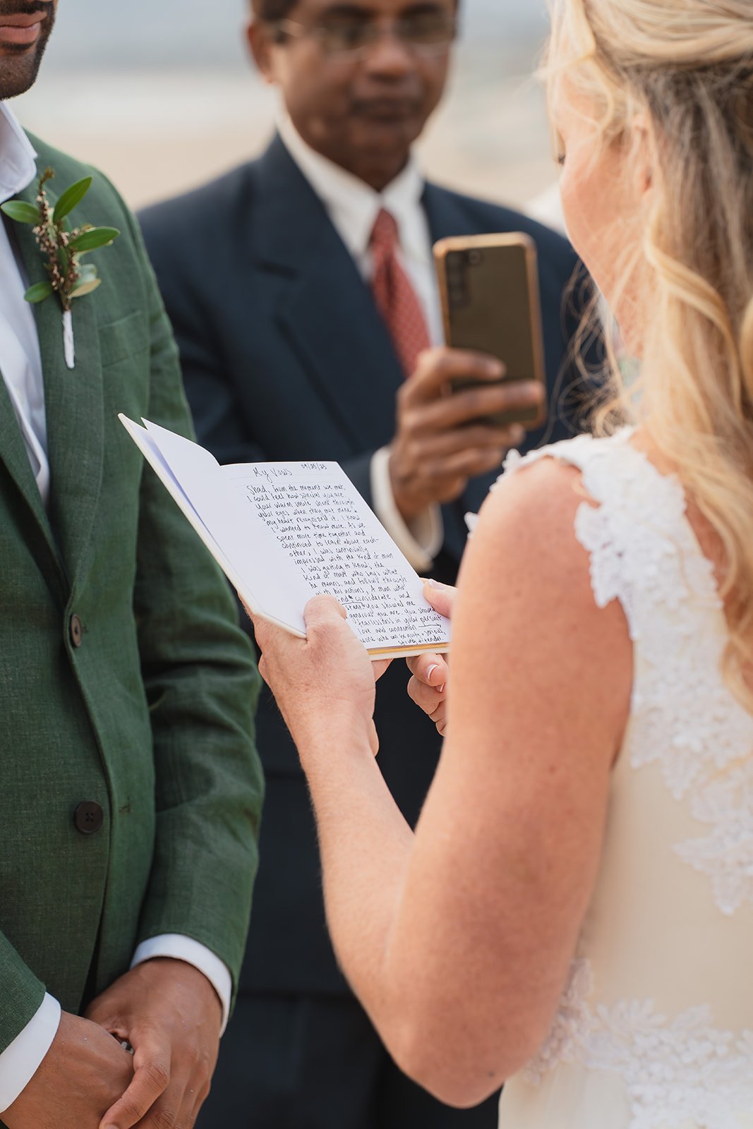 A bride and groom exchange vows during their wedding ceremony, with the bride reading from a small book and the groom listening, while an officiant records the moment with a smartphone.